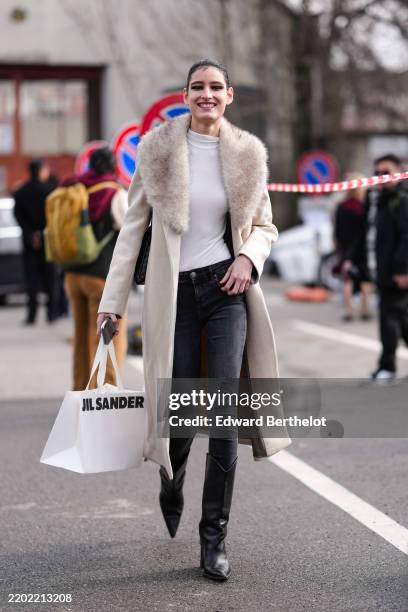 Model wears black eye shadow, white long sleeve shirt, cream wool coat with light gray faux fur collar, a black denim jean skinny pants, shiny black...