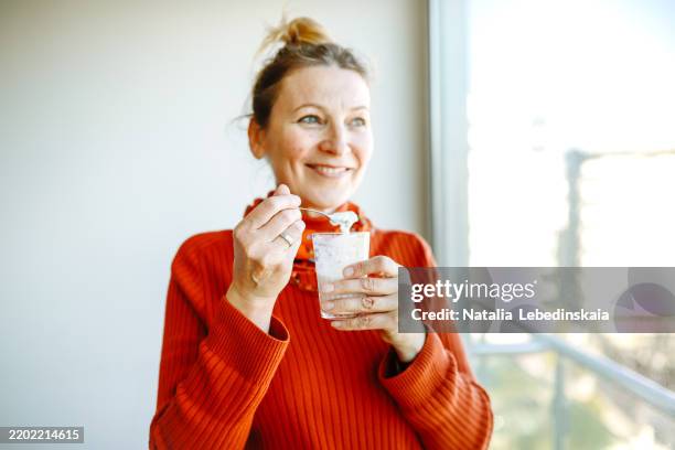 smiling woman in a cozy sweater eats homemade yogurt with granola, basking in the morning sunlight by the window. - produit laitier photos et images de collection