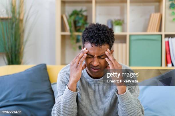 man suffering from headache touching his temples while sitting on sofa at home - headache stock pictures, royalty-free photos & images