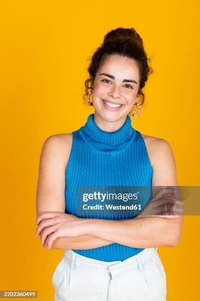 happy young woman standing with arms crossed against yellow background - vestido azul imagens e fotografias de stock