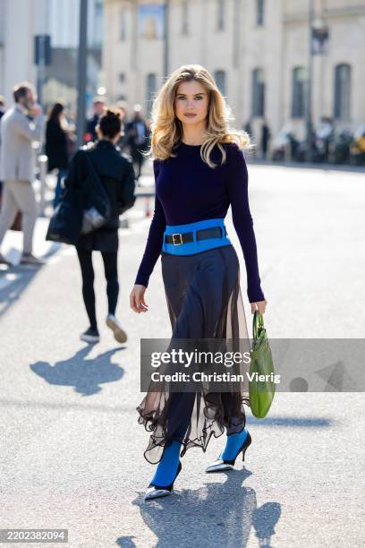 Xenia Adonts wears purple long shirt, high waisted blue transparent brown skirt, blue tights, green bag outside Prada during the Milan Fashion Week...