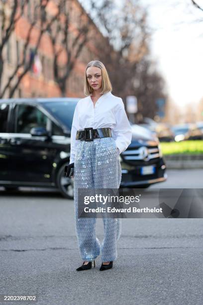 Anouchka Gauthier wears blonde bob hair cut, white oversized buttoned long sleeve shirt, shiny black large leather belt, light blue sequin sheer mesh...