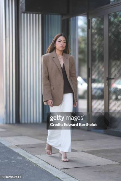 Izabel Sim wears gold earrings, gold layered necklaces, light brown oversized blazer jacket, black v-neck shirt, white draped midi skirt, transparent...