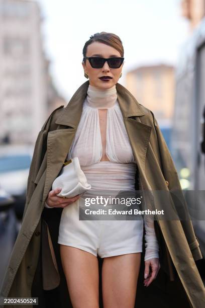 Mary Leest wears black sunglasses, gold earrings, burgundy bordeaux lipstick, white sheer mesh high neck corset top, olive green trench coat, white...