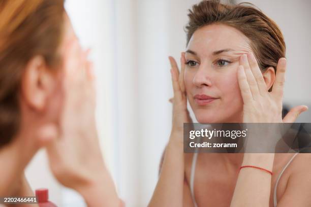 belle femme d’âge moyen massant le visage avec de la crème pour le visage devant le miroir - seulement des femmes dâge moyen photos et images de collection