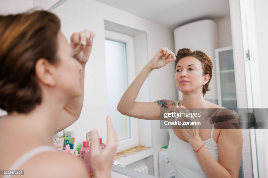 Beautiful mid adult woman tweezing eyebrows in front of the mirror
