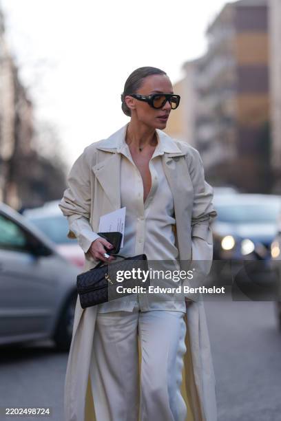 Viktoria Rader wears black sunglasses, silver green Van Cleef & Arpel necklace, cream half buttoned silk long sleeve shirt, beige trench coat, shiny...