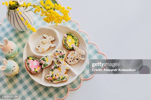 plate with tasty easter cookies in shape on bunny and eggs, mimosa flowers and decoration on light table background. happy easter! - osterkuchen stock-fotos und bilder