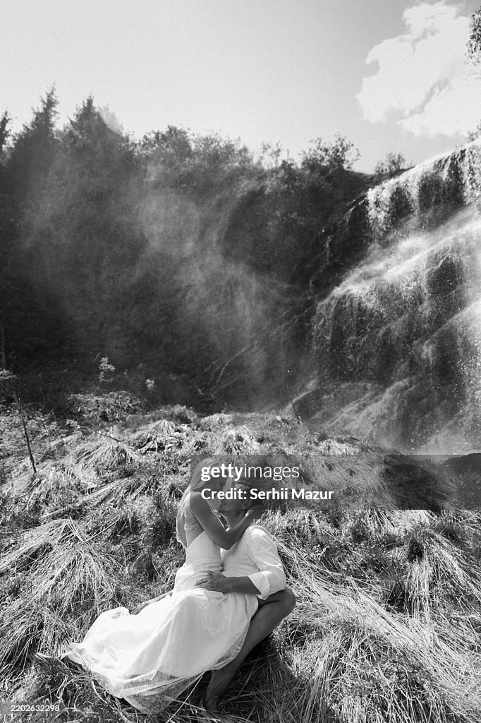 Love-struck newlyweds in festive attire kiss against the backdrop of a waterfall. Black and white stock photo.