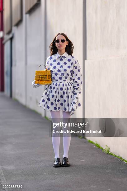 Guest wears black sunglasses, white buttoned up long sleeve dark blue polka dots, white high waisted dark blue polka dotted pleated mini skirt, light...