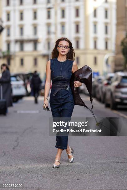 Guest wears a dark blue denim sleeveless dress, a dark brown leather bag, silver shiny pointed shoes, outside Sportmax, during the Milan Fashion Week...