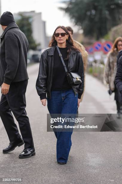 Guest wears brown sunglasses, a black top, a black leather jacket, a black leather bag, blue denim jeans / pants, outside Ferragamo, during the Milan...