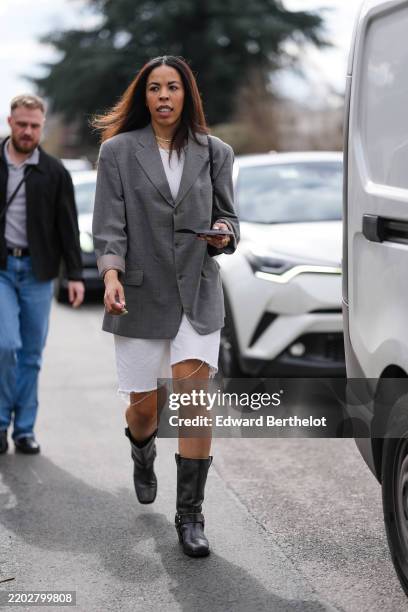 Guest wears gold layered necklace, white t-shirt, dark gray pattern oversized blazer jacket, gold rings, white denim jean shorts, shiny black biker...