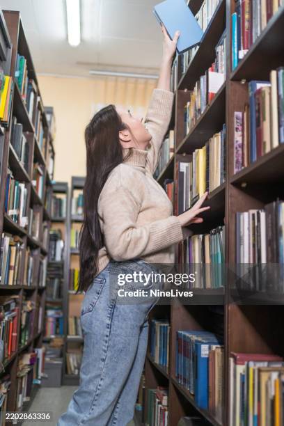 young female student picking a book from library bookshelf - public library stock pictures, royalty-free photos & images
