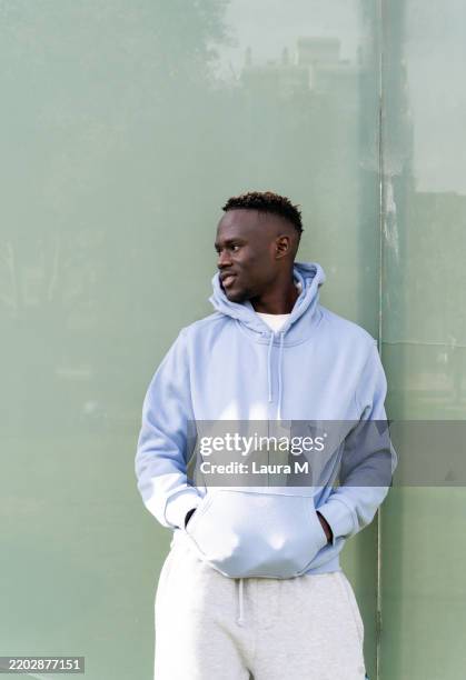 junger mann mit blauem kapuzenpullover und grauer jogginghose schaut in der nähe einer grünen wand weg - kapuzenoberteil stock-fotos und bilder
