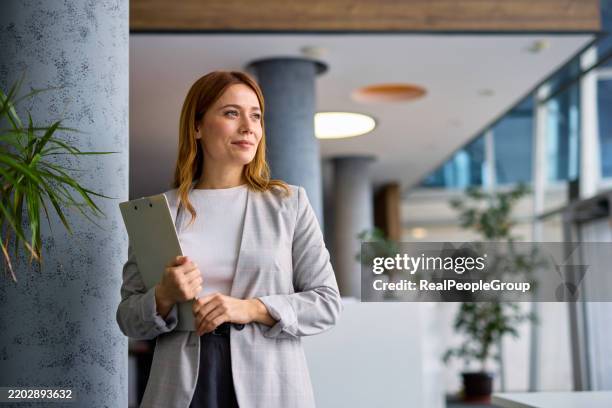 businesswoman holding clipboard looking away in modern office - redhead stock pictures, royalty-free photos & images