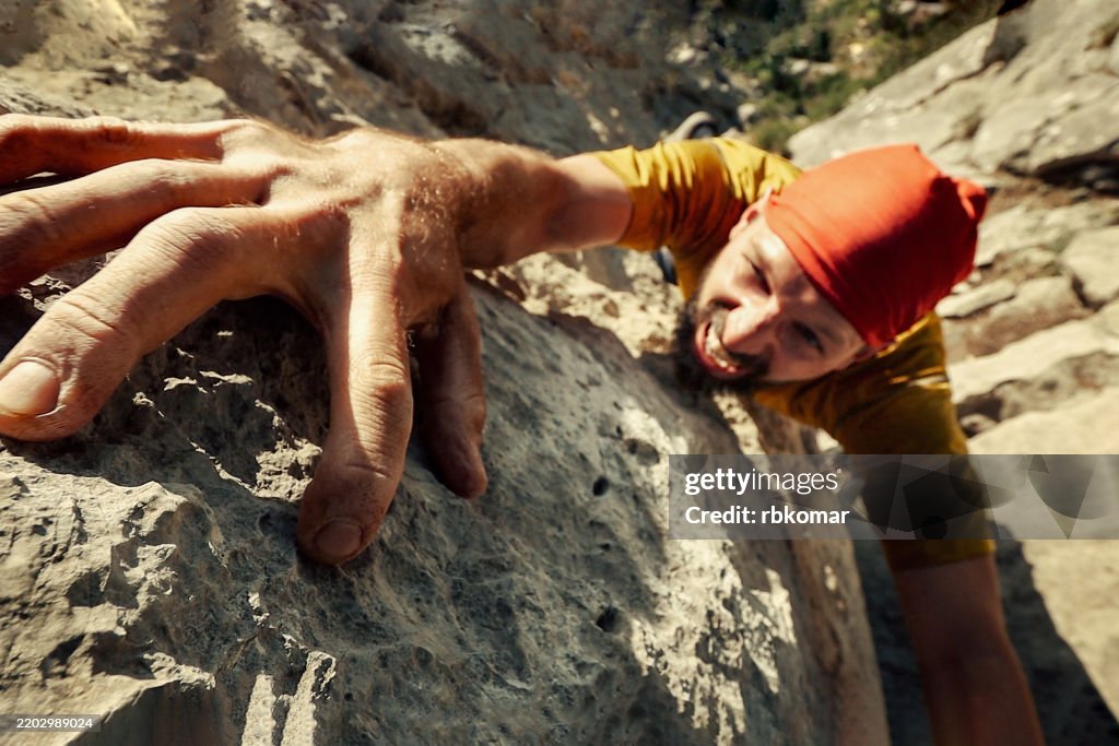 Rock climber grips cliff face during ascent - man with red bandana grimaces with determination while scaling rocky mountain
