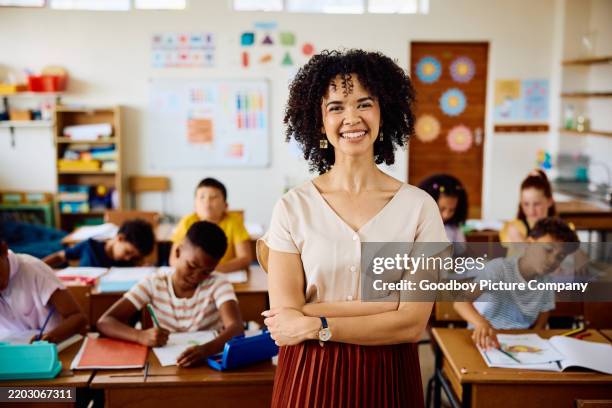 young female teacher with curly hair in classroom, smiling at camera with arms crossed - yngre skolåldern bildbanksfoton och bilder
