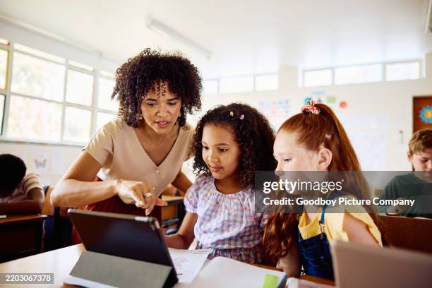 junge lehrerin, die schülerinnen mit einem digitalen tablet anleitet, sitzt am schreibtisch im unterricht - lehrkraft stock-fotos und bilder