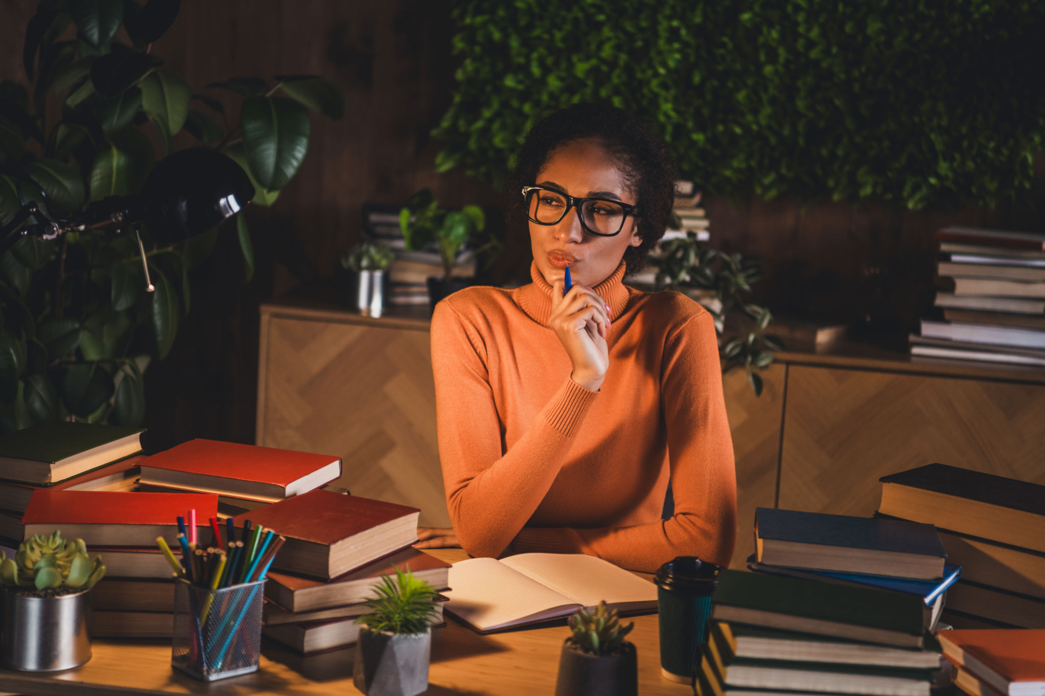 Young woman in cozy attire studies at home desk, surrounded by stacks of books, pens, and green plants Young woman in cozy attire studies at home desk, surrounded by stacks of books, pens, and green plants