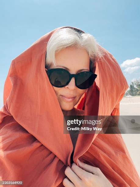 young woman with silver hair wears oversized sunglasses and a burnt orange hooded scarf. beach setting and bright blue sky provide a striking contrast to her bold fashion choice. - high fashion stock pictures, royalty-free photos & images