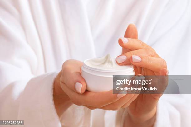 beautiful woman in white bathrobe holds white jar with beauty product moisturizing facial cream or hand cream with swirl top. - retinol stock pictures, royalty-free photos & images