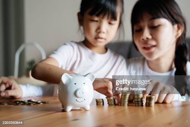 asian mother and daughter putting coins into piggy bank - welvaart stockfoto's en -beelden