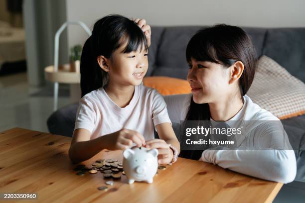 asian mother and daughter putting coins into piggy bank - welvaart stockfoto's en -beelden