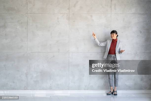 smiling business woman presenting on a concrete wall space - teacher pointing finger stock pictures, royalty-free photos & images