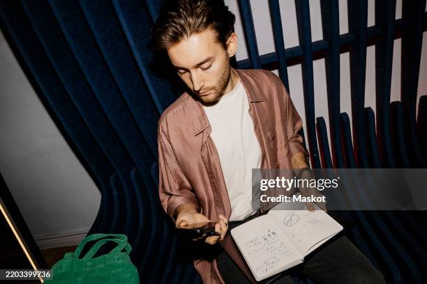 high angle view of male entrepreneur using smart phone and holding diary while sitting on seat at office - phone flash light stock pictures, royalty-free photos & images