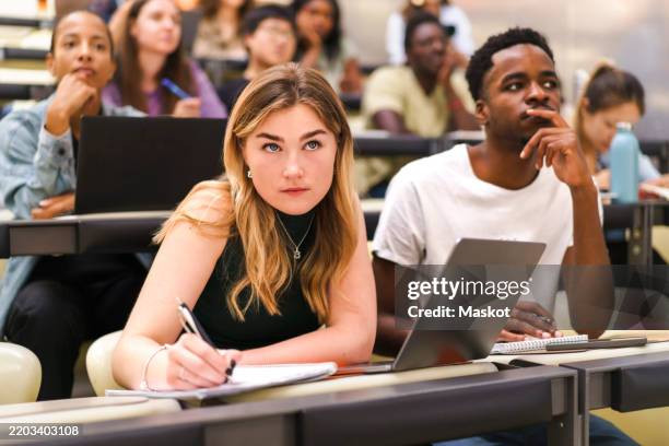 young female student writing in book while sitting by male friend at university classroom - sala de aula de universidade imagens e fotografias de stock