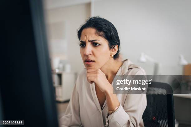 confused female creative professional using desktop pc while sitting with hand on chin in office - enredado fotografías e imágenes de stock