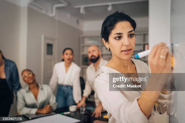 female entrepreneur writing on whiteboard during meeting with coworkers in office - plano de atividades imagens e fotografias de stock