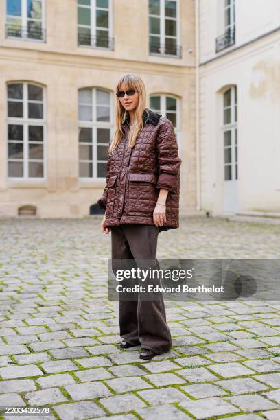 Guest wears dark brown sunglasses, shiny burgundy bordeaux quilted oversized jacket, a gold bracelet, gold rings, dark brown loose denim jean pants,...
