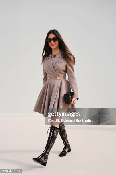 Guest wears black sunglasses, a gold necklace, a gold ring, light brown double breasted blazer jacket midi dress, shiny black belted leather bag,...