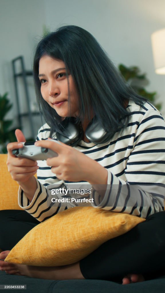 Young Asian woman looking at camera using wireless controller playing console game to win sit on sofa at living room at home