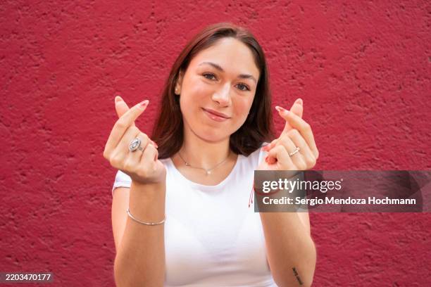 young latino woman smiling, looking at the camera and making a heart gesture with her hands, love - hand sign stock pictures, royalty-free photos & images