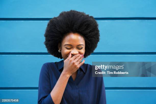 woman laughing, hand over her mouth, next to blue wall - verlegen stockfoto's en -beelden