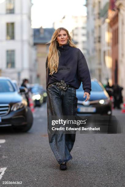 Elle Ferguson wears dark gray half zipped high neck Acne Studios sweater, shiny black leather belt with silver buckle detail, dark gray loose baggy...
