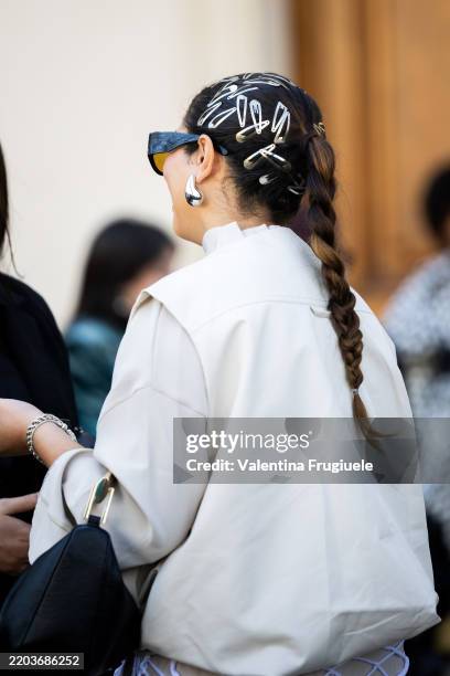 Guest wears silver hairpins, silver drop-shaped earrings, black sunglasses, a black bag and a white jacket outside Rabanne show during the Womenswear...