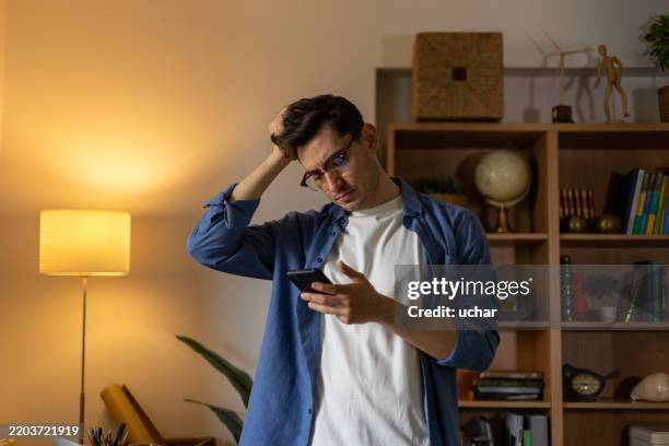 young man scratching his head while reading upsetting news on his smartphone - verward stockfoto's en -beelden