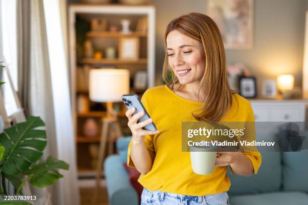 woman smiling while holding coffee and using smartphone - telefonia imagens e fotografias de stock