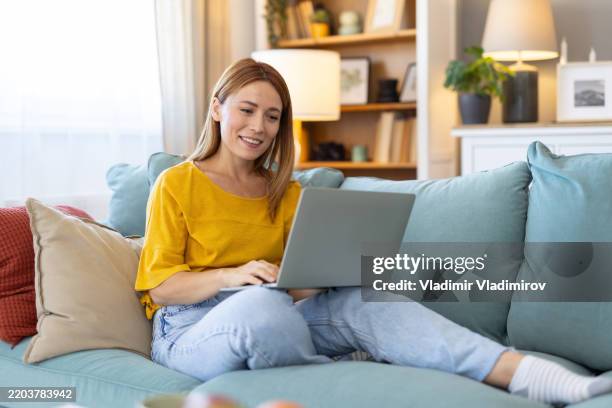 smiling woman using laptop in a bright living room - financial literacy stock pictures, royalty-free photos & images