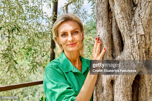 smiling blond mature woman hugging olive tree and looking at camera - abbracciare le ginocchia foto e immagini stock