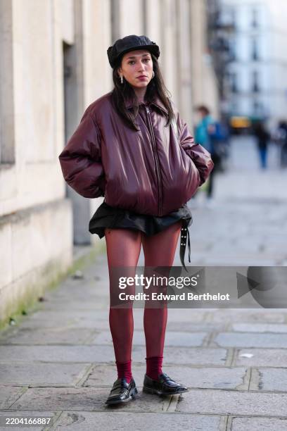 Guest wears black oversized baseball cap hat, silver hoop earrings, shiny burgundy bordeaux zipped bomber leather jacket, black mini skirt, burgundy...