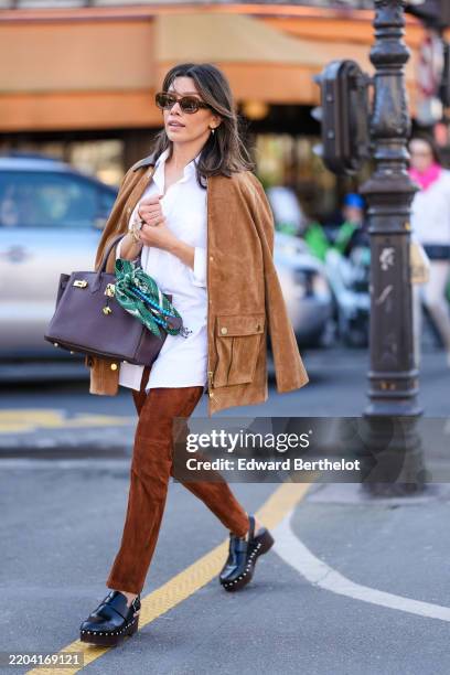 Guest wears dark brown sunglasses, gold earrings, white buttoned up long sleeve oversized shirt, light brown oversized suede jacket, shiny dark brown...