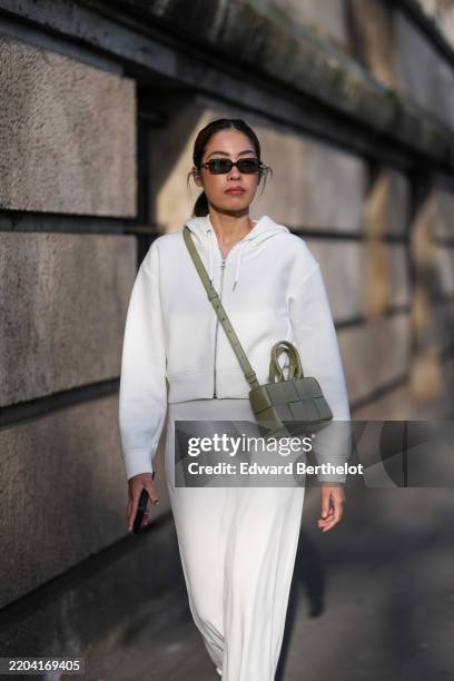 Izabel Sim wears black sunglasses, white zipped hoodie, white loose pants, dark olive green woven leather bag, outside Elie Saab, during the Paris...