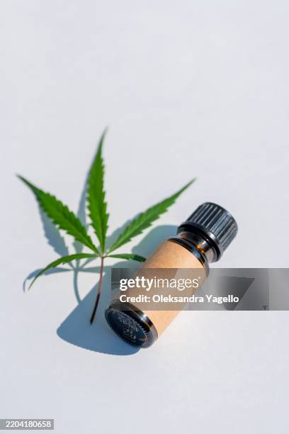 natural hemp extract bottle alongside a green cannabis leaf on a white surface during daylight - hemp agriculture stock pictures, royalty-free photos & images
