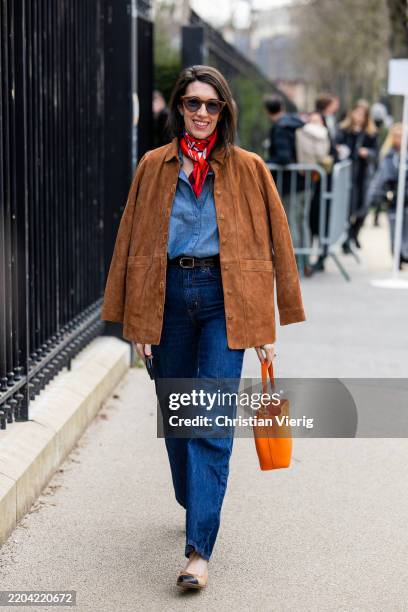Guest wears brown suede jacket, navy blue denim jeans, orange bag, blouse, red scarf outside Lacoste during the Womenswear Fall/Winter 2025/2026 as...