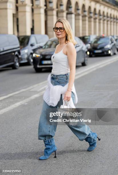 Iryna Thater wears white Loewe shirt, jeans, denim boots & wrapped pullover Loewe, sunglasses YSL, white Jacquemus bag during the Womenswear...